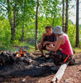 Henkilöitä nuotion ääressä kesäisellä rannalla juomassa nokipannukahvia.