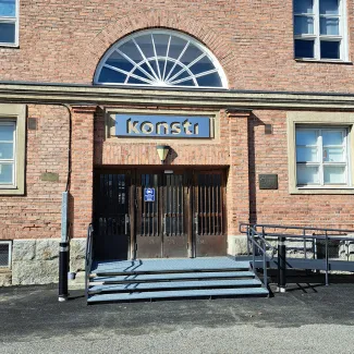 Punatiilinen rakennus, lähikuva ulko-ovesta. Oven yläpuolella puolipyöreä ikkuna ja kyltti, jossa lukee konsti