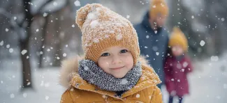 Lapsi hymyilee talvivaatteissa ulkona, lunta sataa ja taustalla muitakin ihmisiä.